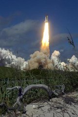 Space Shuttle Atlantis roars into the sky above the primitive sand dunes in the foreground.