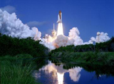 Launch of Space Shuttle Atlantis into the deep blue sky is reflected in the nearby water framed by the green underbrush. Clouds of smoke and steam spread side to side across Launch Pad 39B.