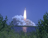 Space Shuttle Atlantis hurtles toward space, propelled by twin columns of fire from the solid rocket boosters, which are captured in the reflection on the water.